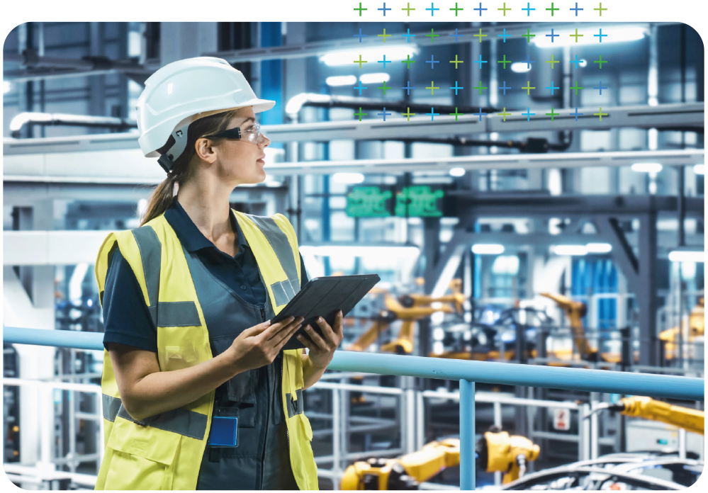 a woman in a hard hat and safety vest is holding a tablet in a factory.