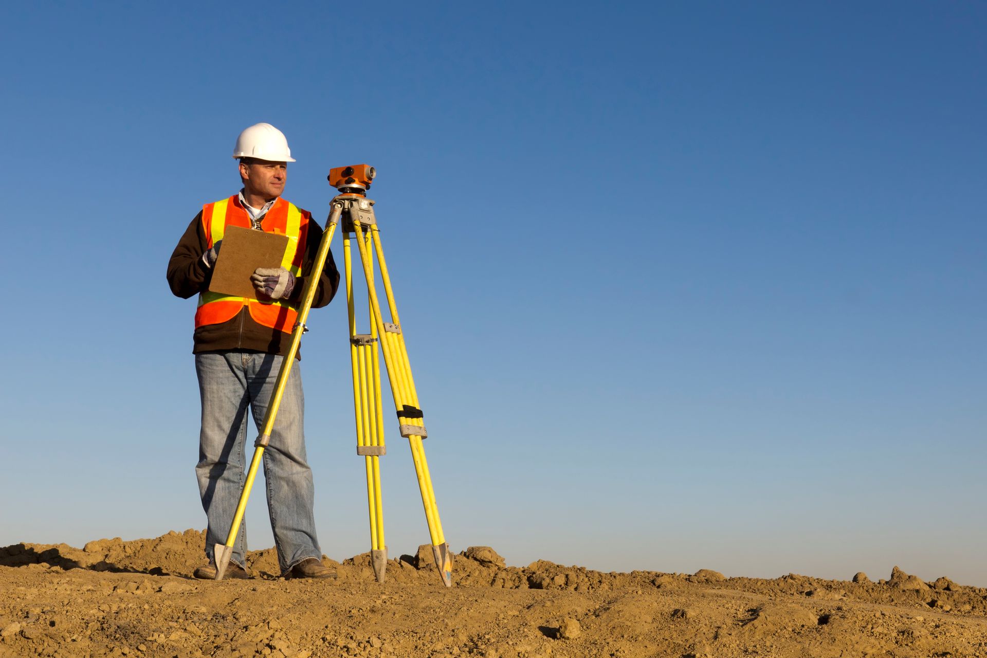 A man is standing in the dirt with a clipboard and a tripod.