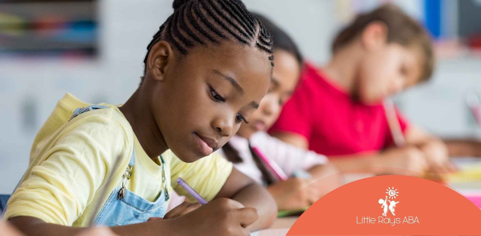A young Black girl writing at a desk with other students in a classroom.