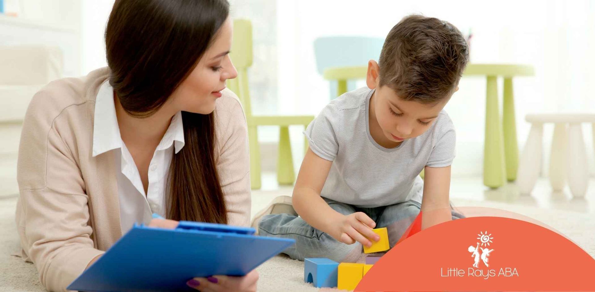 Woman with clipboard observes a boy playing with blocks on the floor, in a room with colorful furniture.