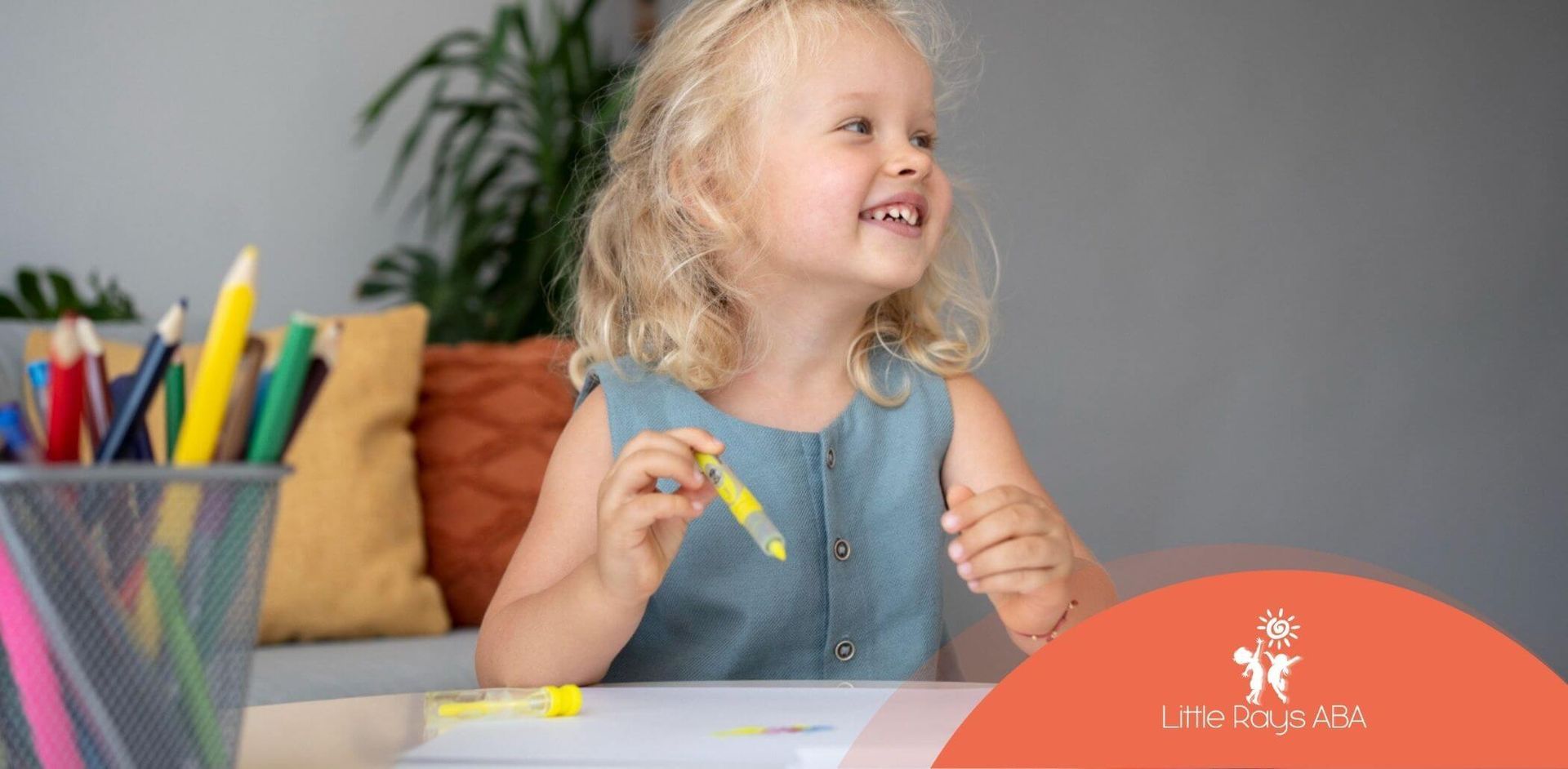 A young, blond girl smiles while holding a yellow marker, sitting at a table with art supplies.