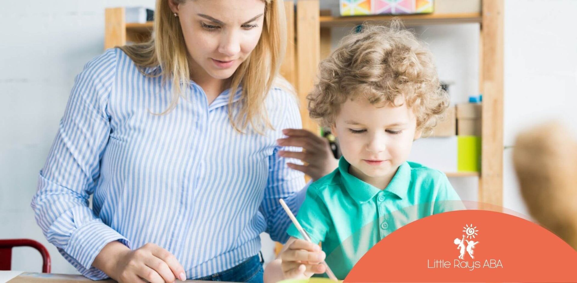 An ABA therapist helps a young boy paint on paper at a table with watercolors in ABA therapy.
