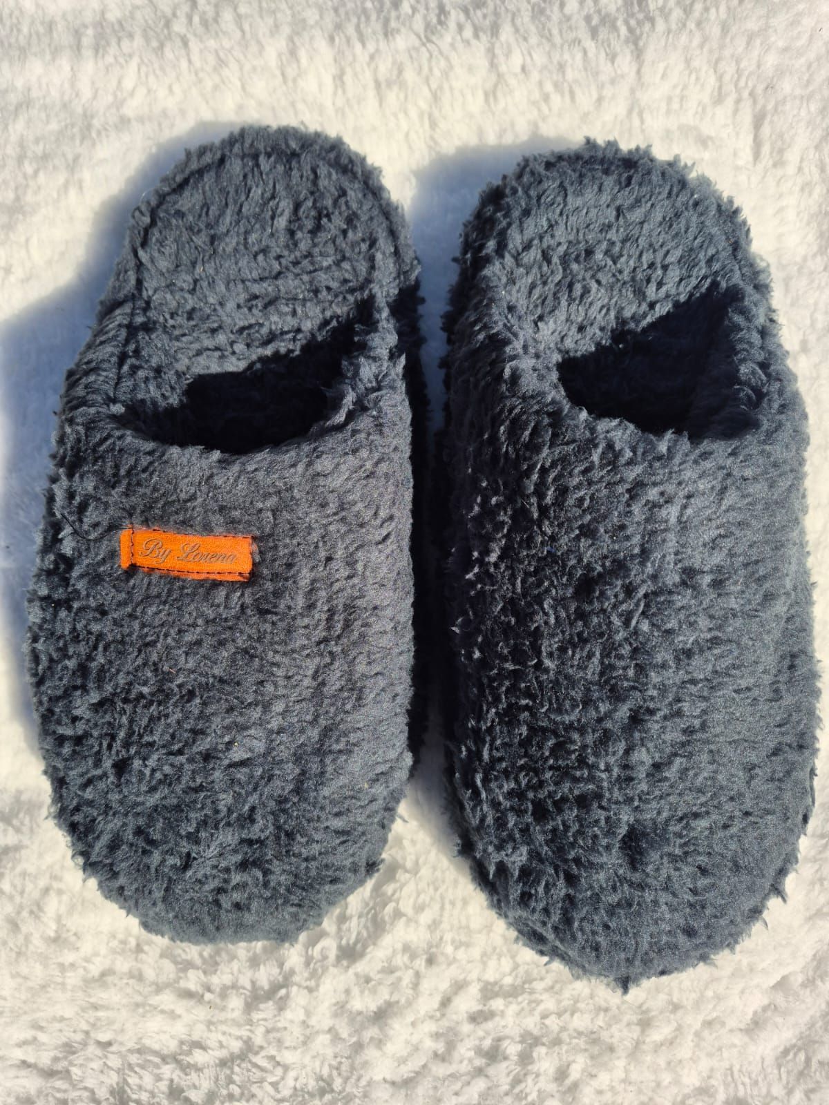 Par de pantuflas de felpa gris oscuro sobre una superficie de textura clara, una de ellas con una pequeña etiqueta naranja.