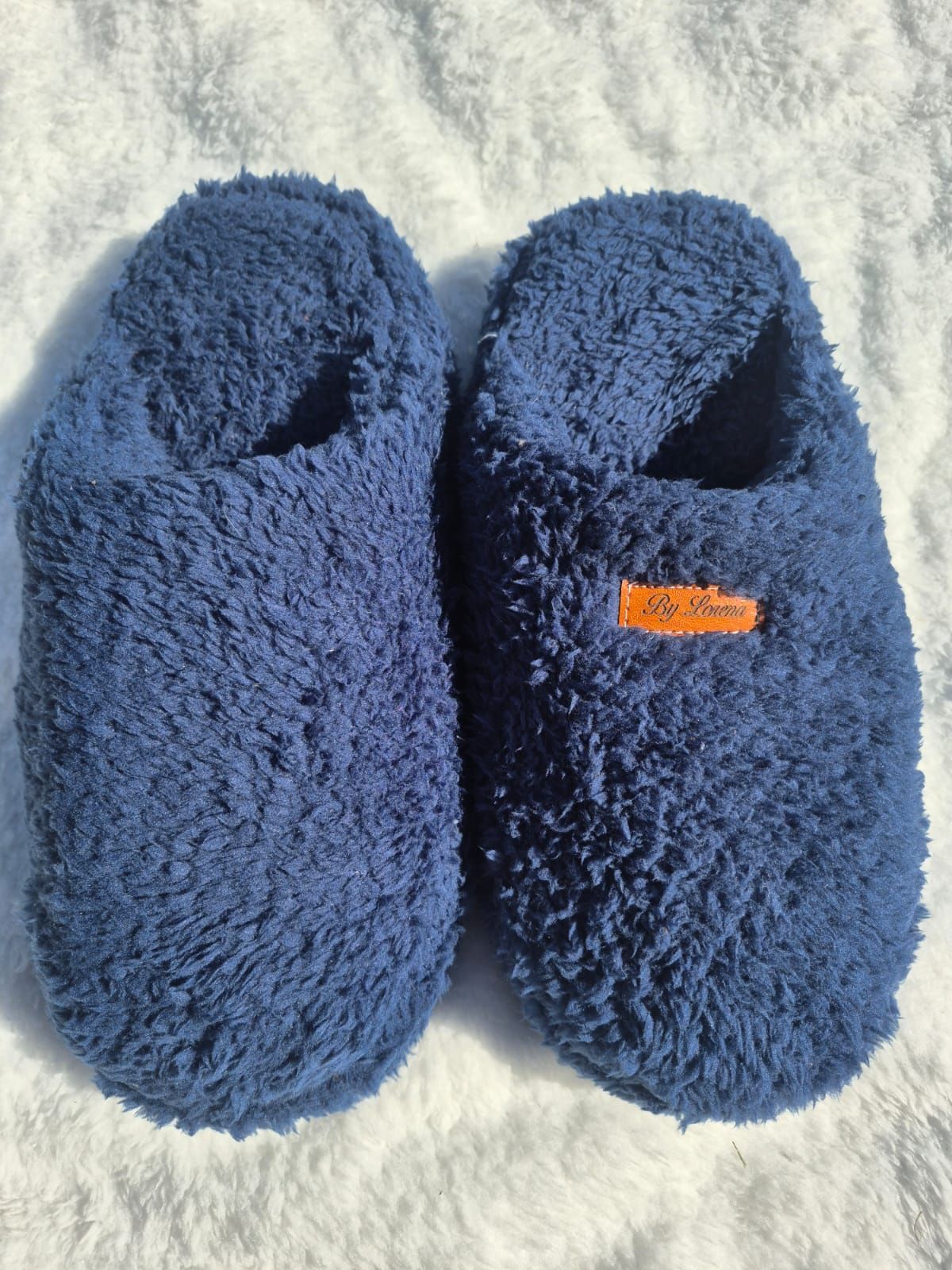 Un par de pantuflas azules de felpa sobre una superficie de textura ligera, una de ellas con una etiqueta naranja.