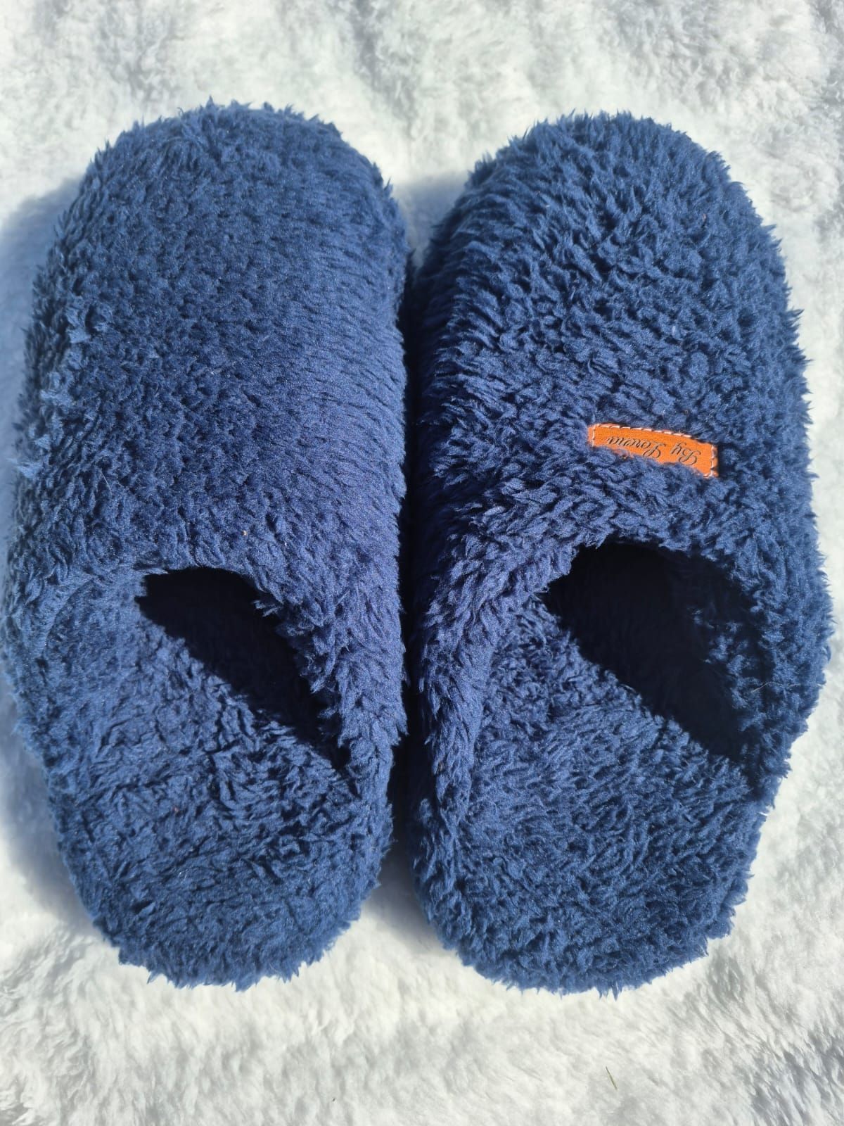 Un par de pantuflas azules y peludas sobre una superficie blanca, una de ellas con una pequeña etiqueta naranja.