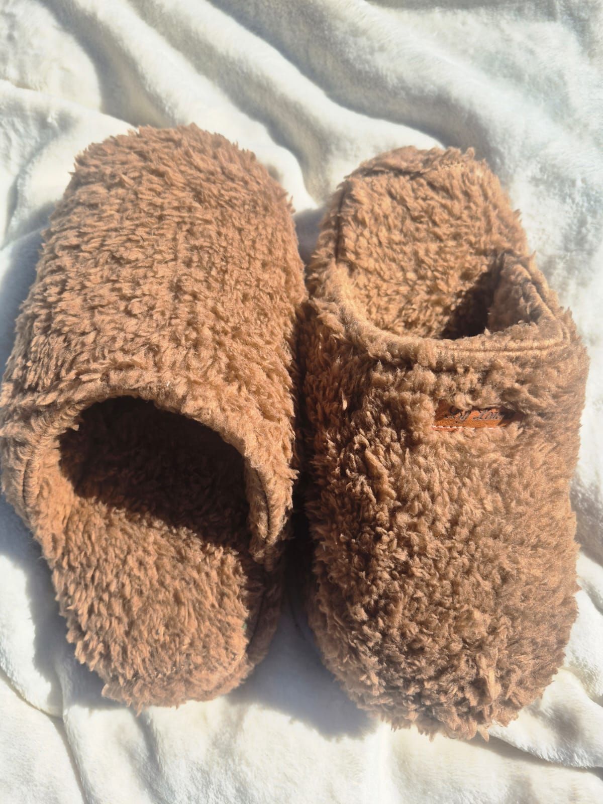Pantuflas marrones y peludas sobre una manta blanca.