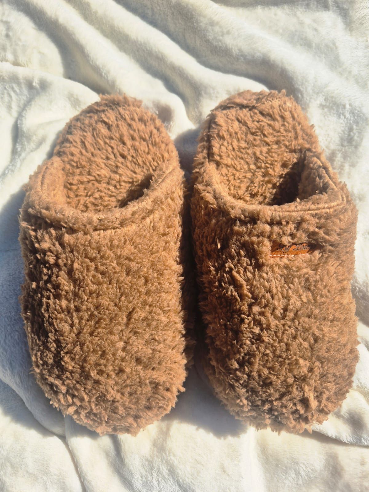 Pantuflas marrones y peludas sobre una manta blanca.