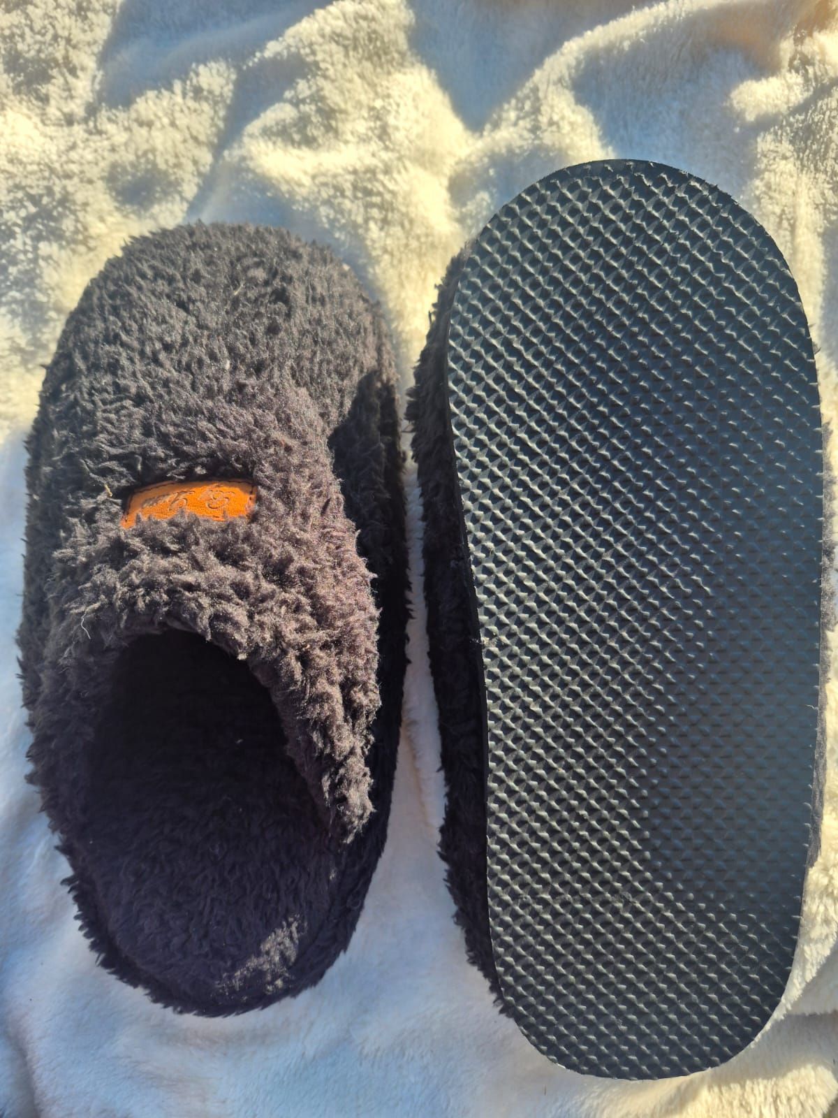 Un par de pantuflas negras de felpa, una de ellas con suela negra de relieve texturizado.