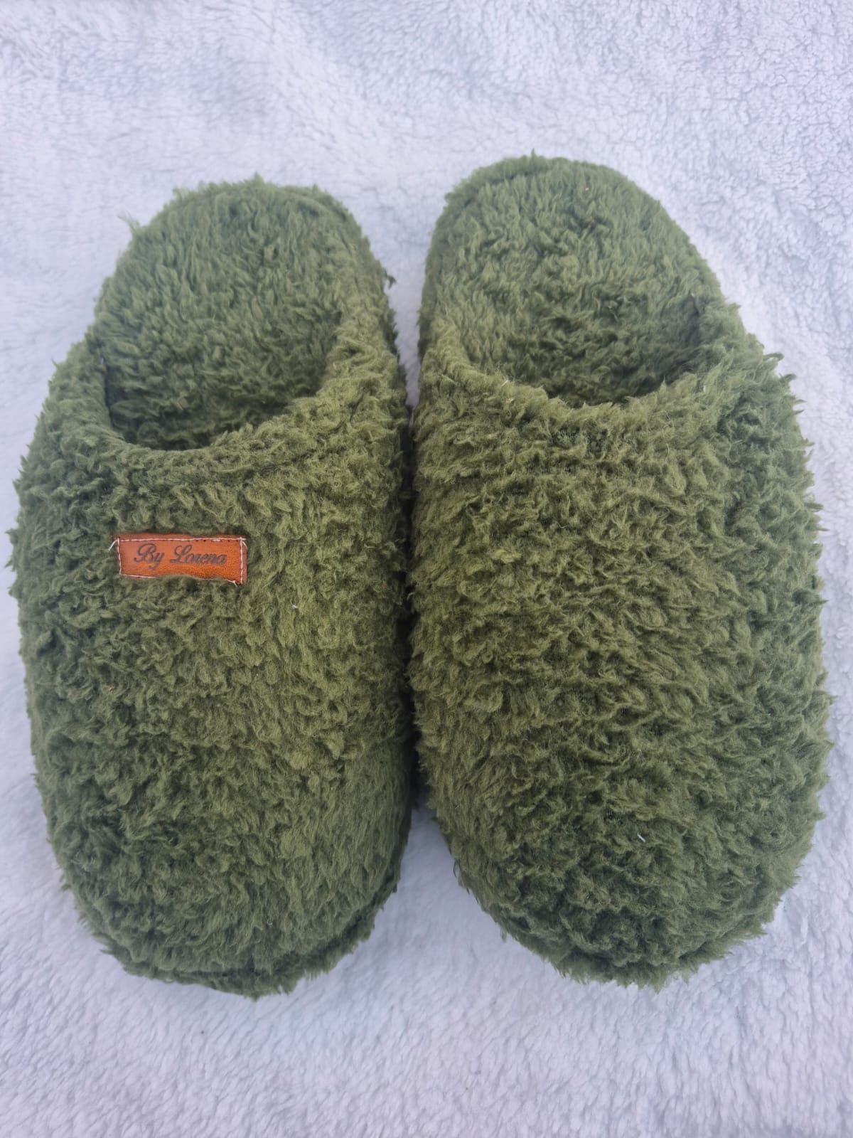 Un par de pantuflas verdes de felpa sin cordones sobre un fondo de toalla de color lila claro.