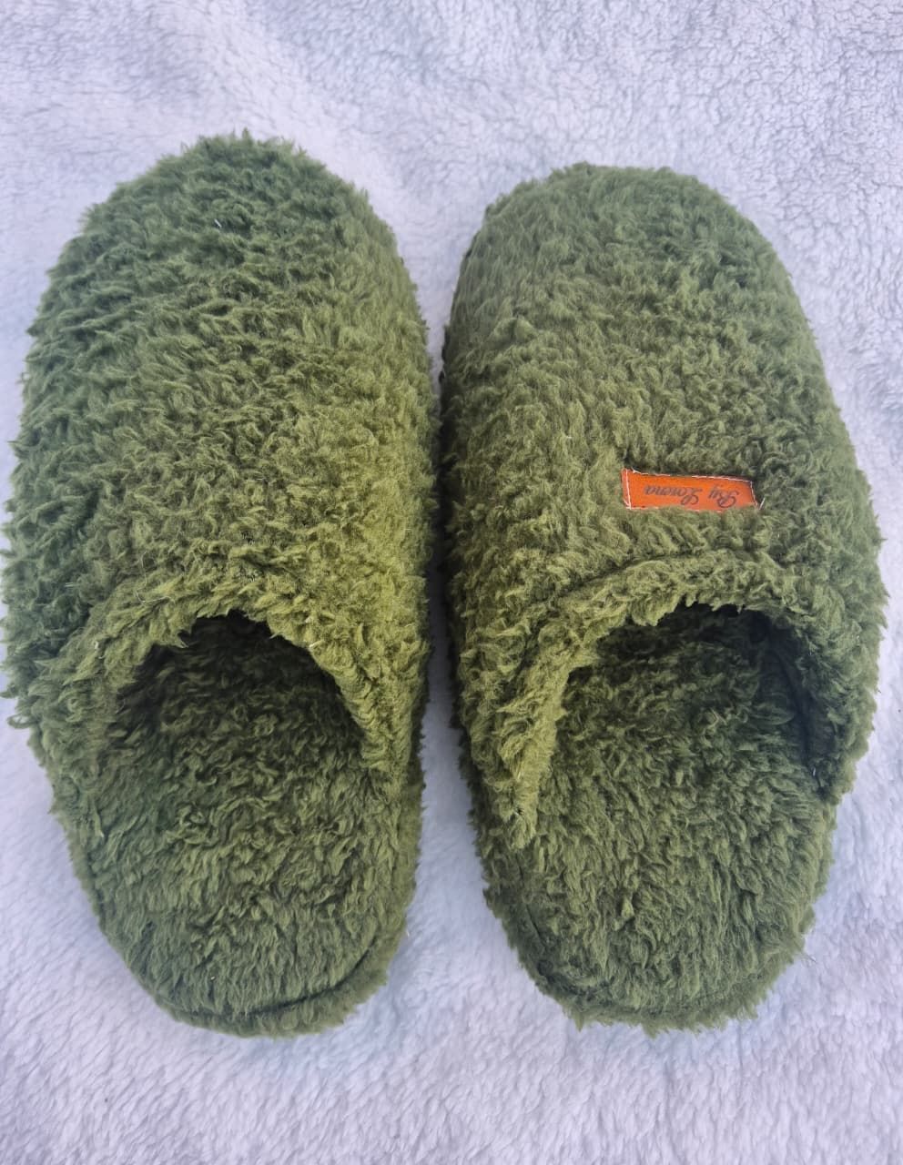 Un par de pantuflas verdes de felpa sin cordones sobre un fondo de toalla de color lila claro.