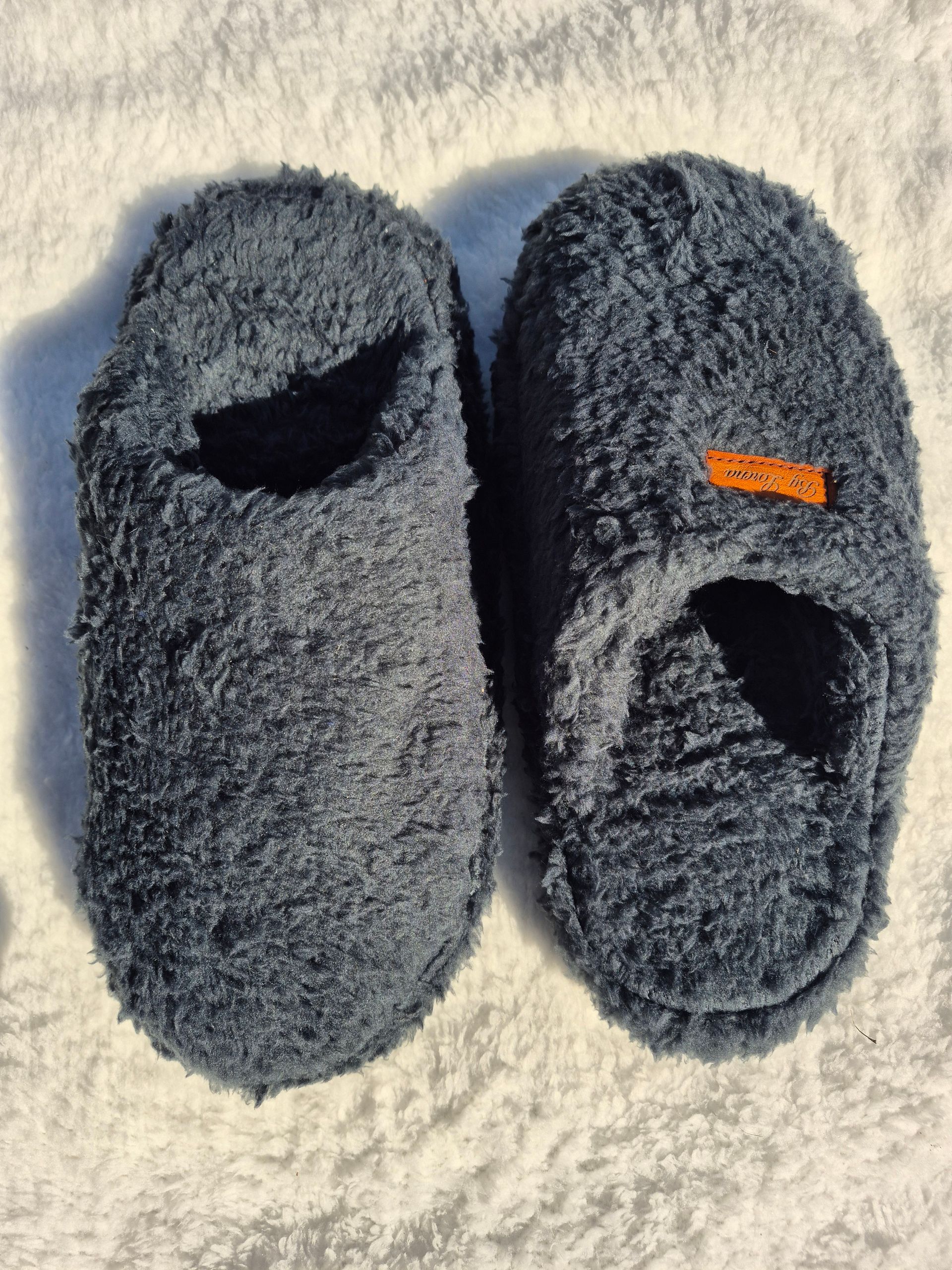 Par de pantuflas de felpa gris oscuro sobre una superficie de textura clara, una de ellas con una pequeña etiqueta naranja.