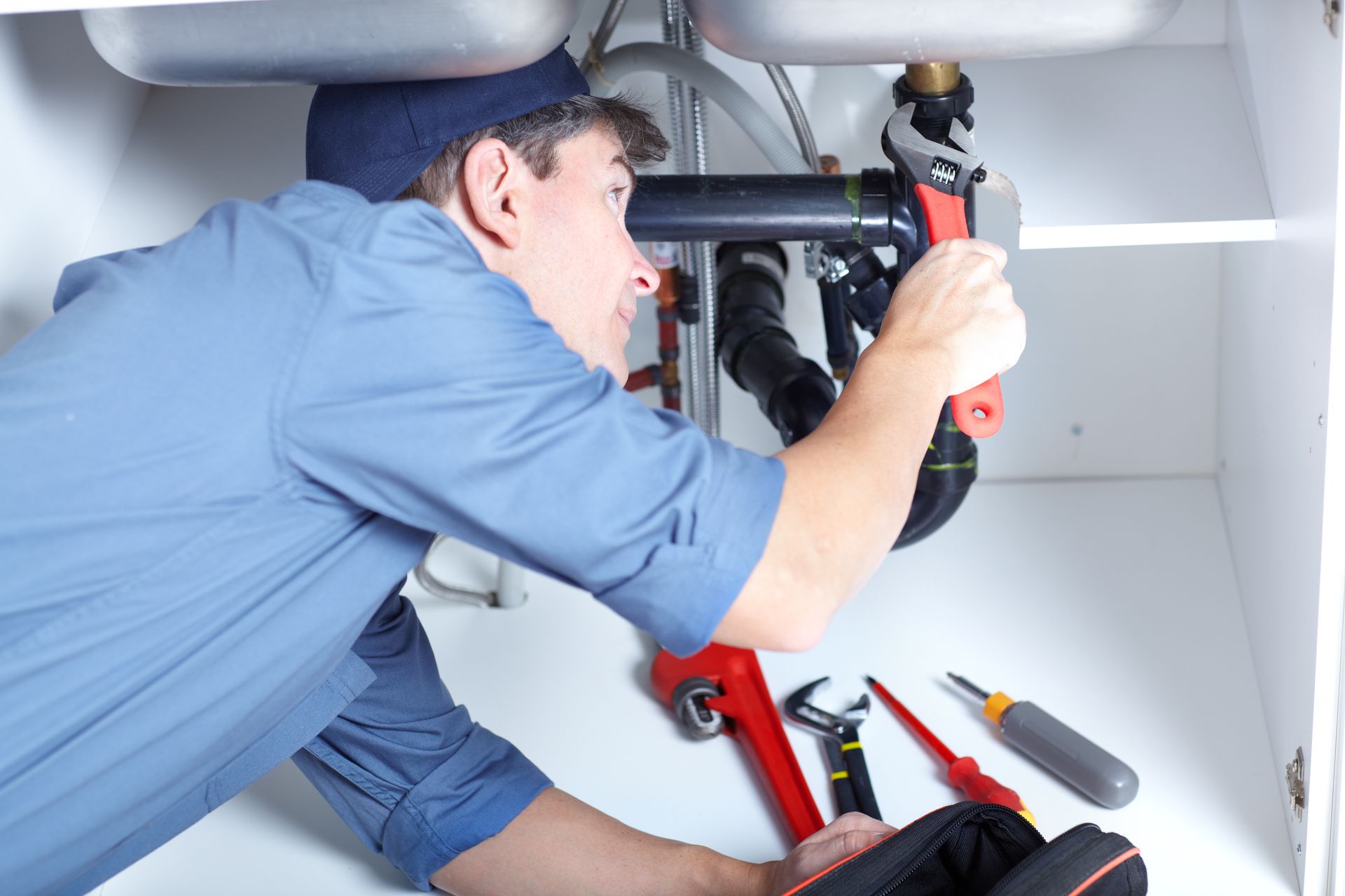 A plumber in a blue uniform tightening a sink pipe with a wrench, tools scattered nearby.