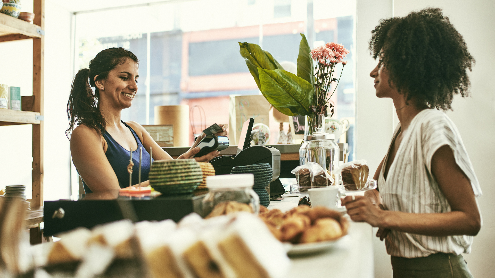 Two women are standing at a counter in a bakery talking to each other.