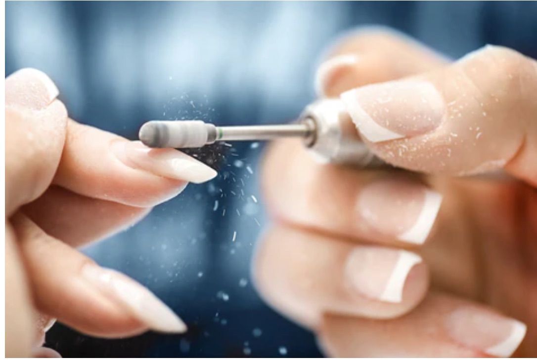 Hands with French-tip nails being filed with a nail drill. Dust particles are visible in the air.