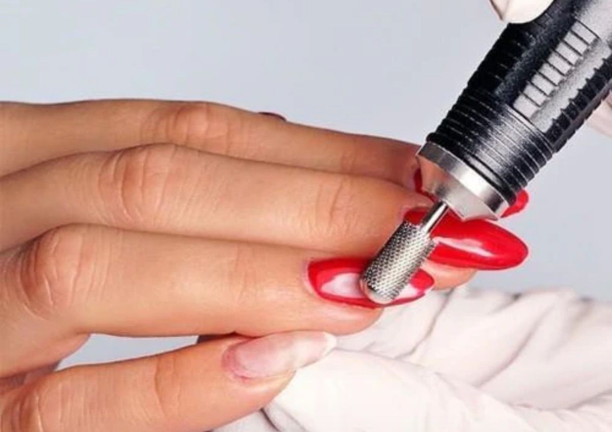 A person filing a red acrylic nail with an electric nail drill, close-up of hand and tool.