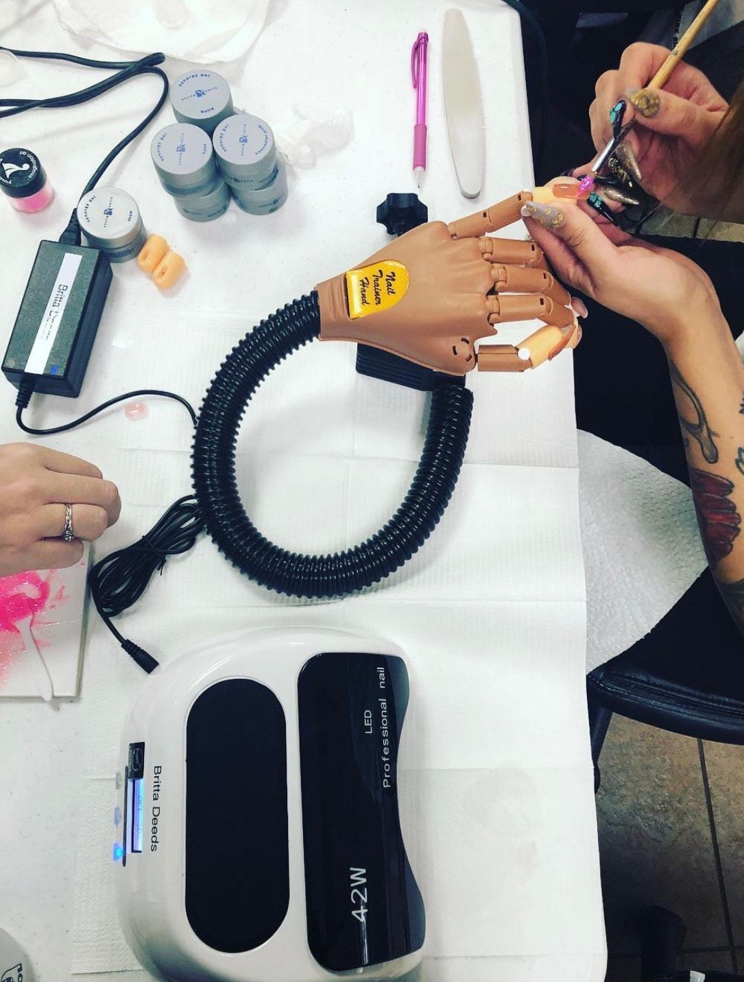 Manicurist working on a prosthetic hand attached to a stand, using tools and gel nail curing lamp.
