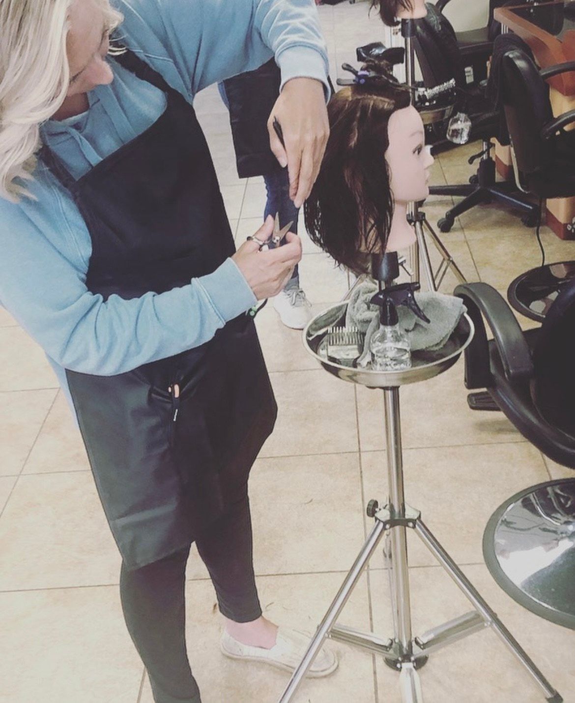 Hairdresser cuts the hair of a mannequin in a salon. She wears a black apron and blue top, with a stand holding tools.
