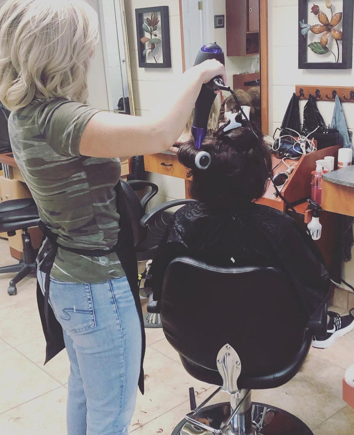 A hair stylist with light blonde hair blow-dries the dark hair of a seated client at a salon.