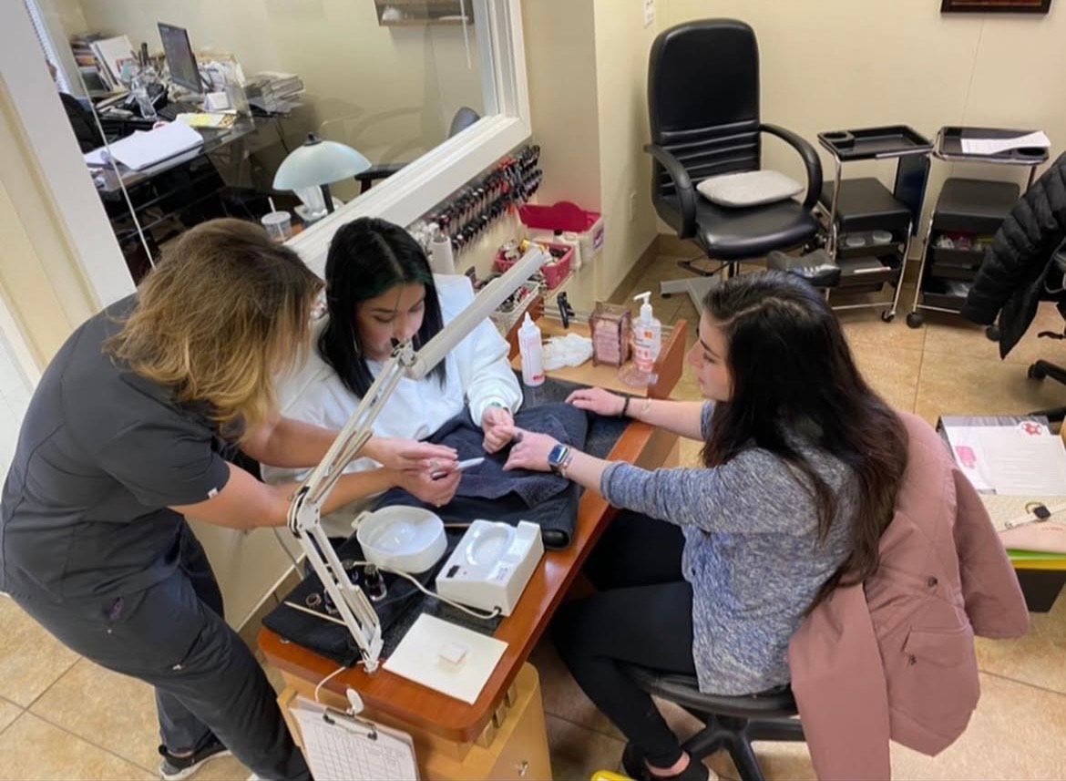 Three women at a nail salon: a nail technician works on a client's feet, while another client has her nails done.