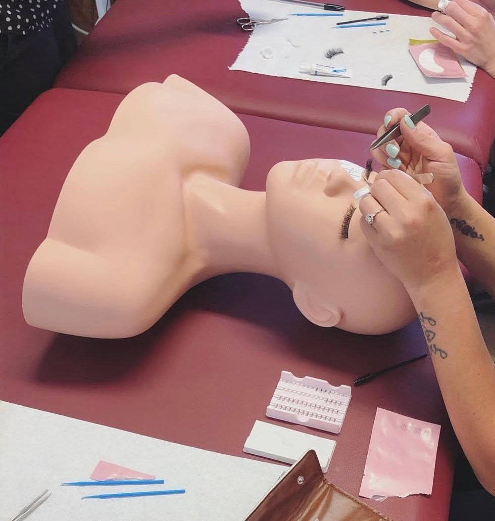 A person practices applying eyelash extensions to a mannequin head on a red table.