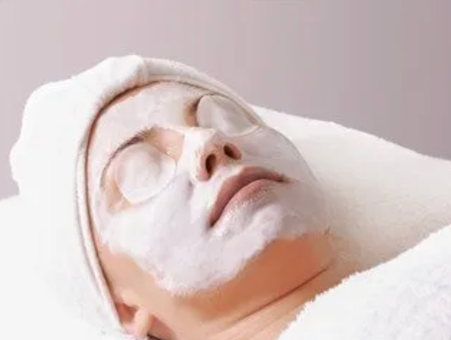 Woman lying down with a white facial mask and towel on her head, in a spa setting.