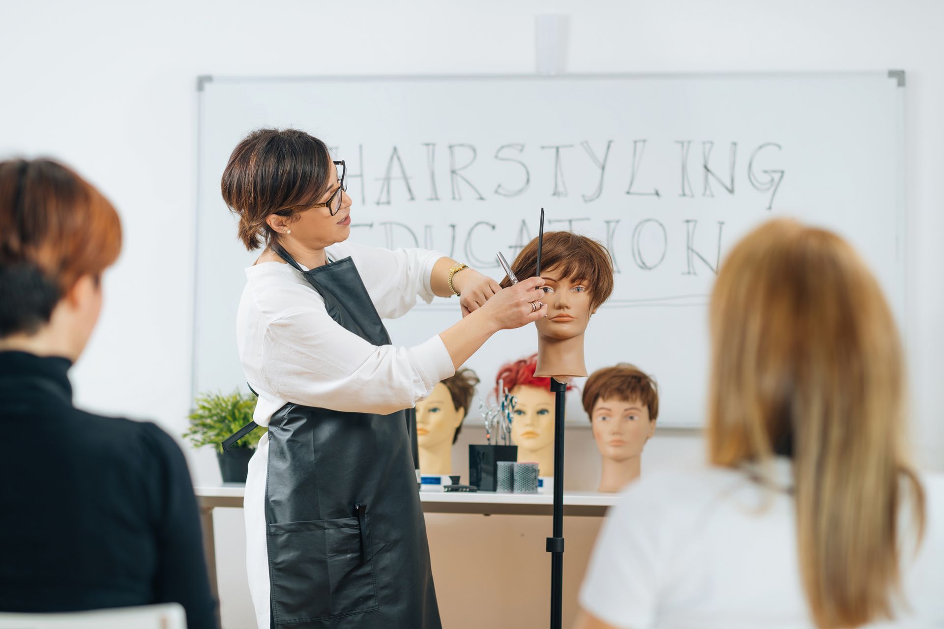 A woman is teaching a group of people how to cut a wig on a mannequin.