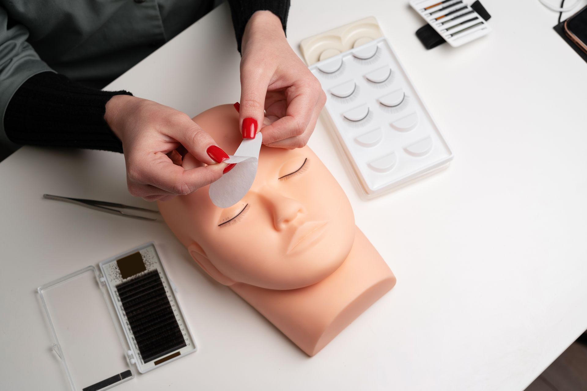 Person applying an eye pad to a mannequin head, preparing for eyelash extensions. Various lash supplies are on the table.
