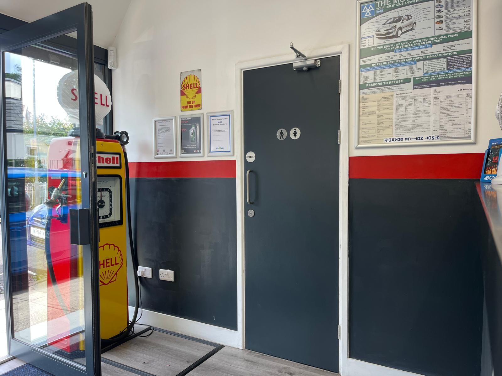 Entrance area with Shell gas pump, black door, red trim, and posters on the wall.