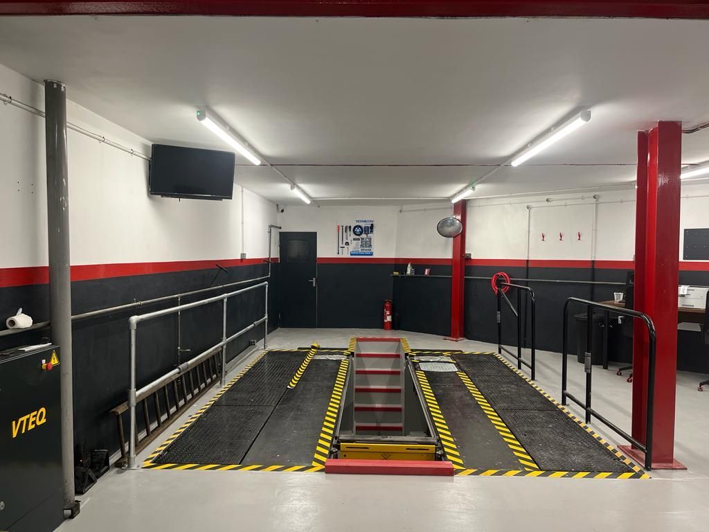 Interior of an auto repair shop with a vehicle lift. Red and black accents, fluorescent lighting, and a TV are visible.