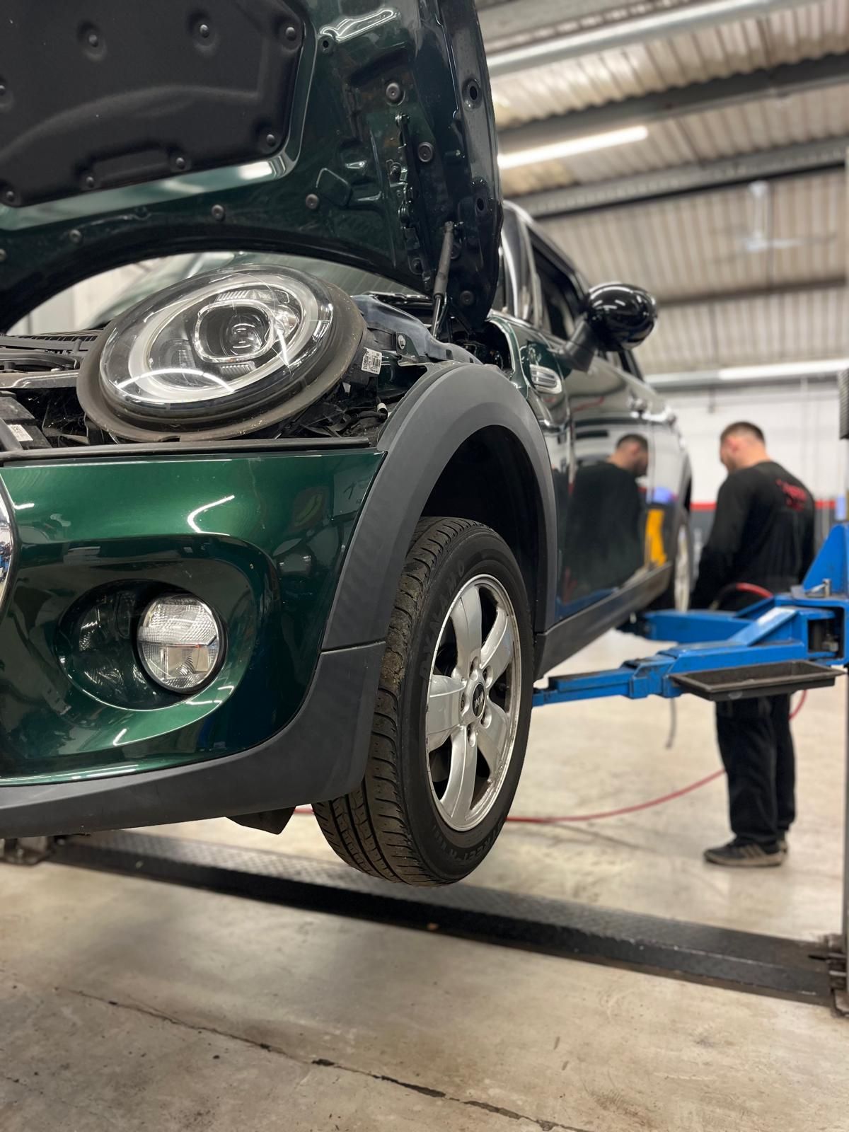 Green Mini Cooper on a lift in a garage with a mechanic observing. Hood open.