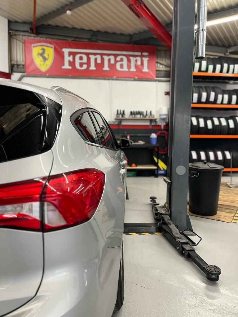 Silver car in a garage with a Ferrari sign and tire rack. Car is near a lift.