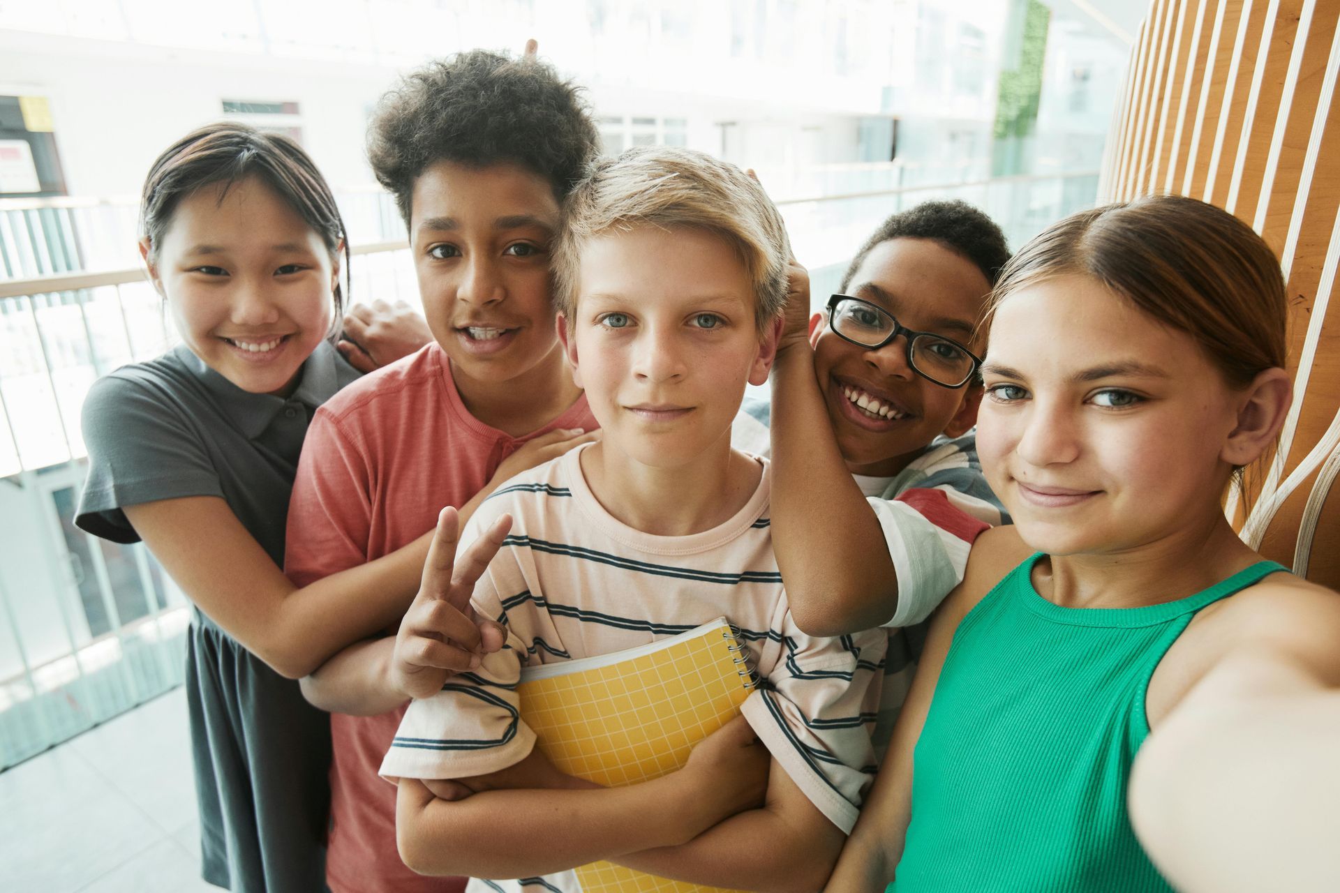 Group of five diverse children smiling for a selfie indoors near a railing.