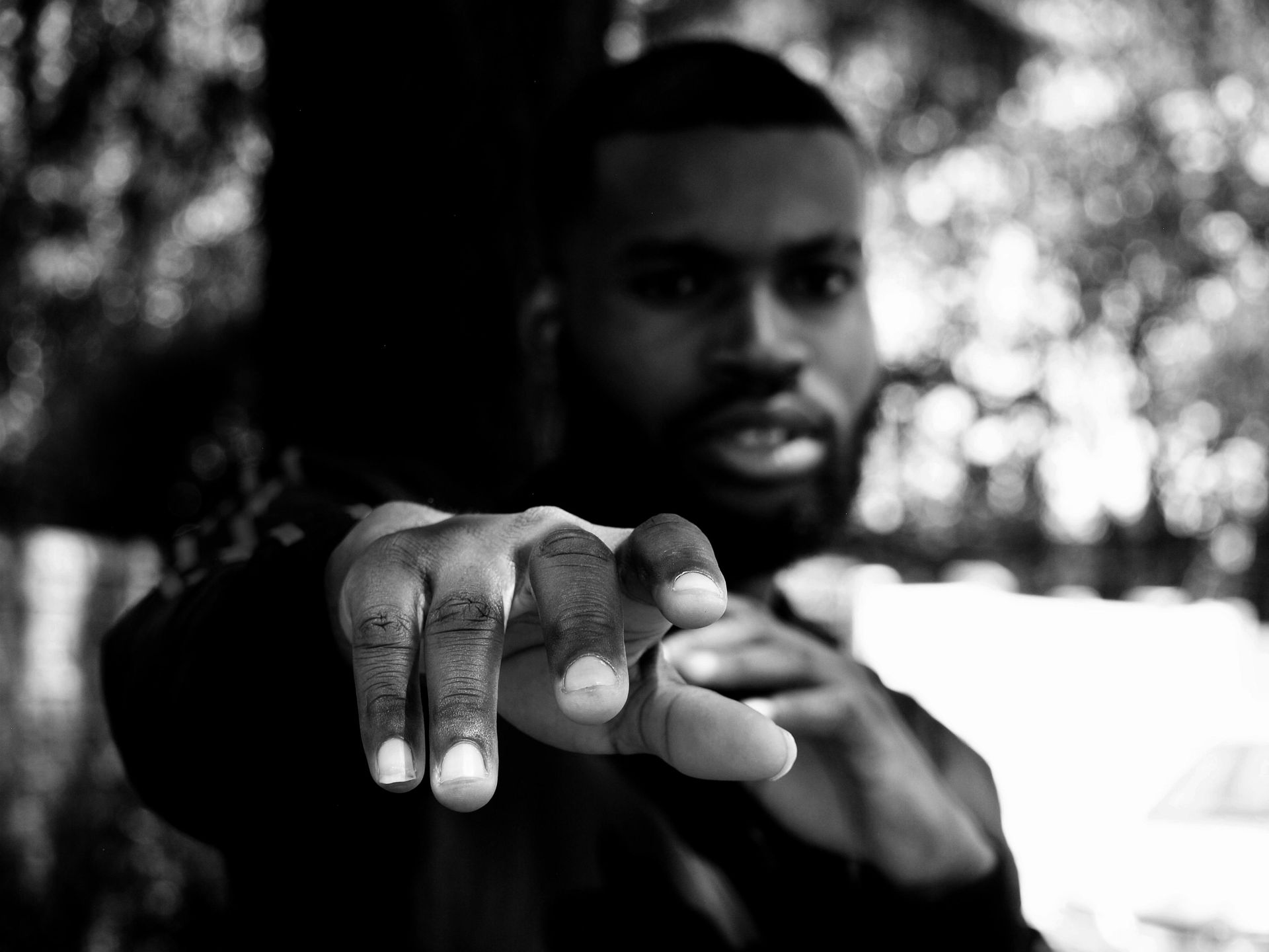 Black and white photo: man reaching forward, fingers extended. Outdoors, blurry background.