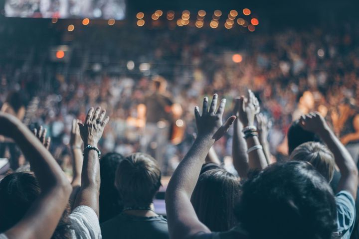 Crowd at a concert with raised hands; stage lights illuminate the band in the background.