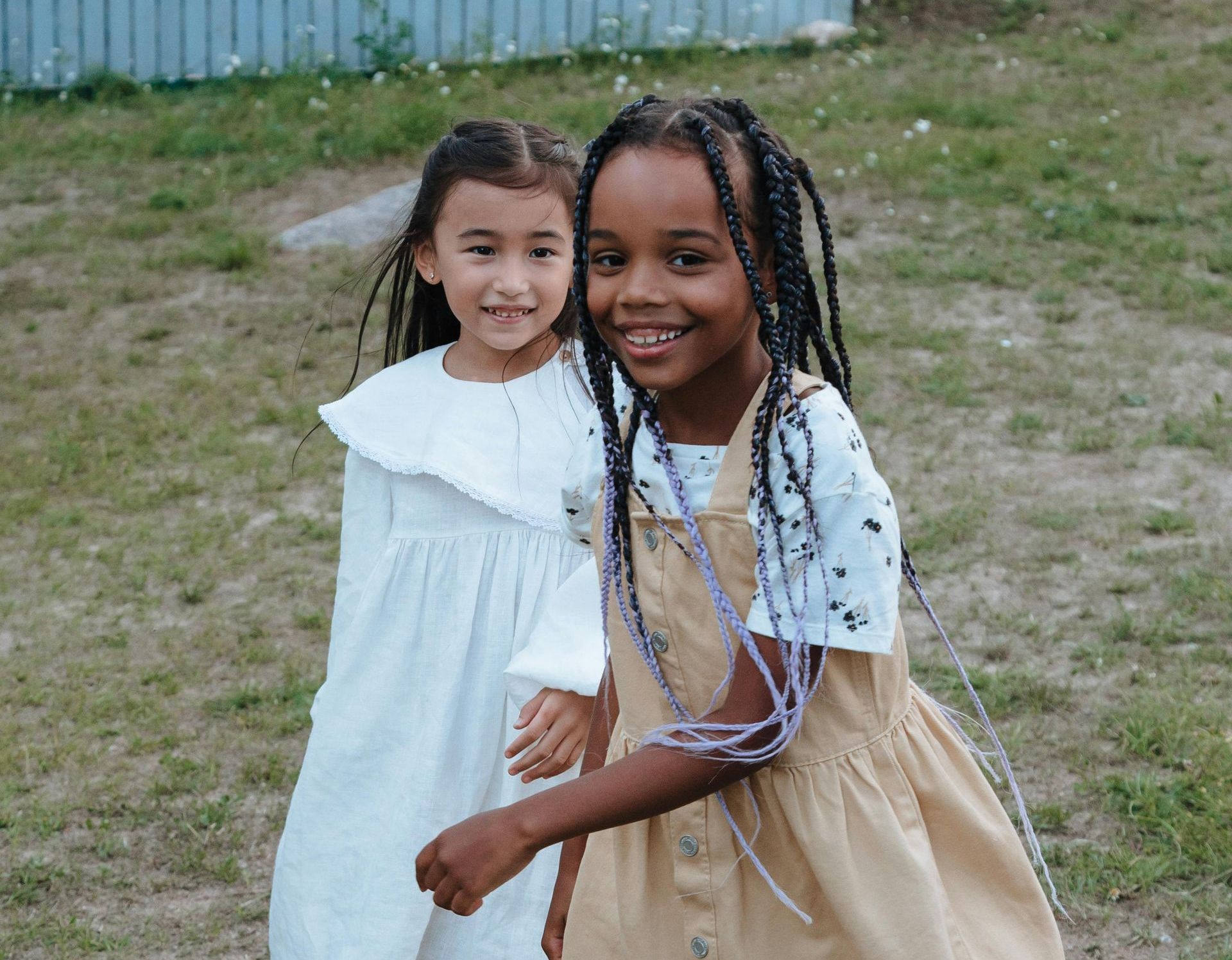 Two young girls smiling outdoors, one in a tan overall dress, the other in a white dress.