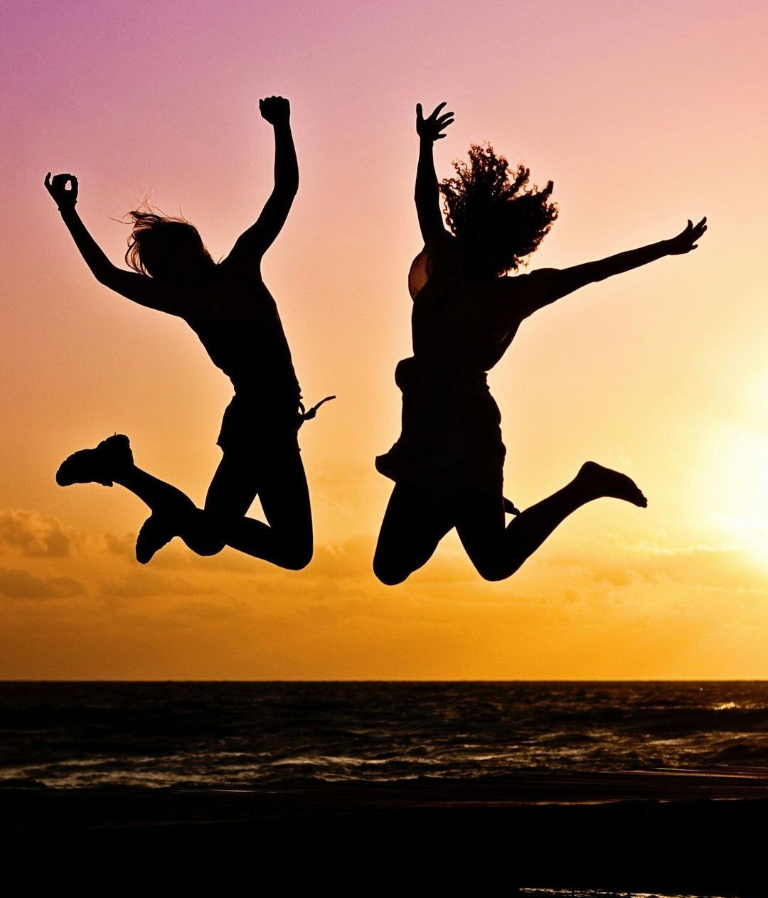 Two people jumping with joy silhouetted against a sunset over the ocean.