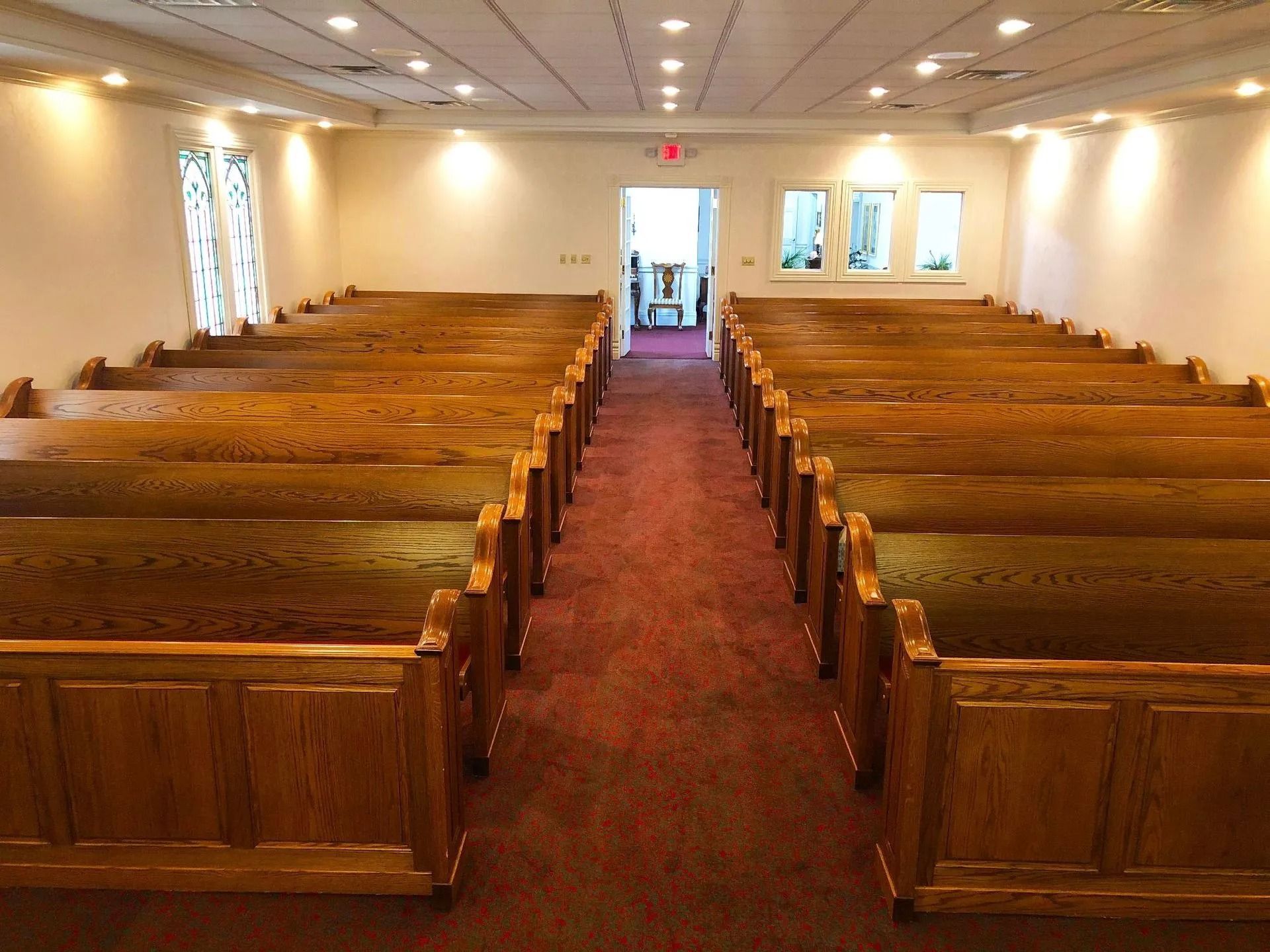 Rows of wooden pews face a center aisle in a church or chapel. Red carpet, cream walls, and recessed lighting.