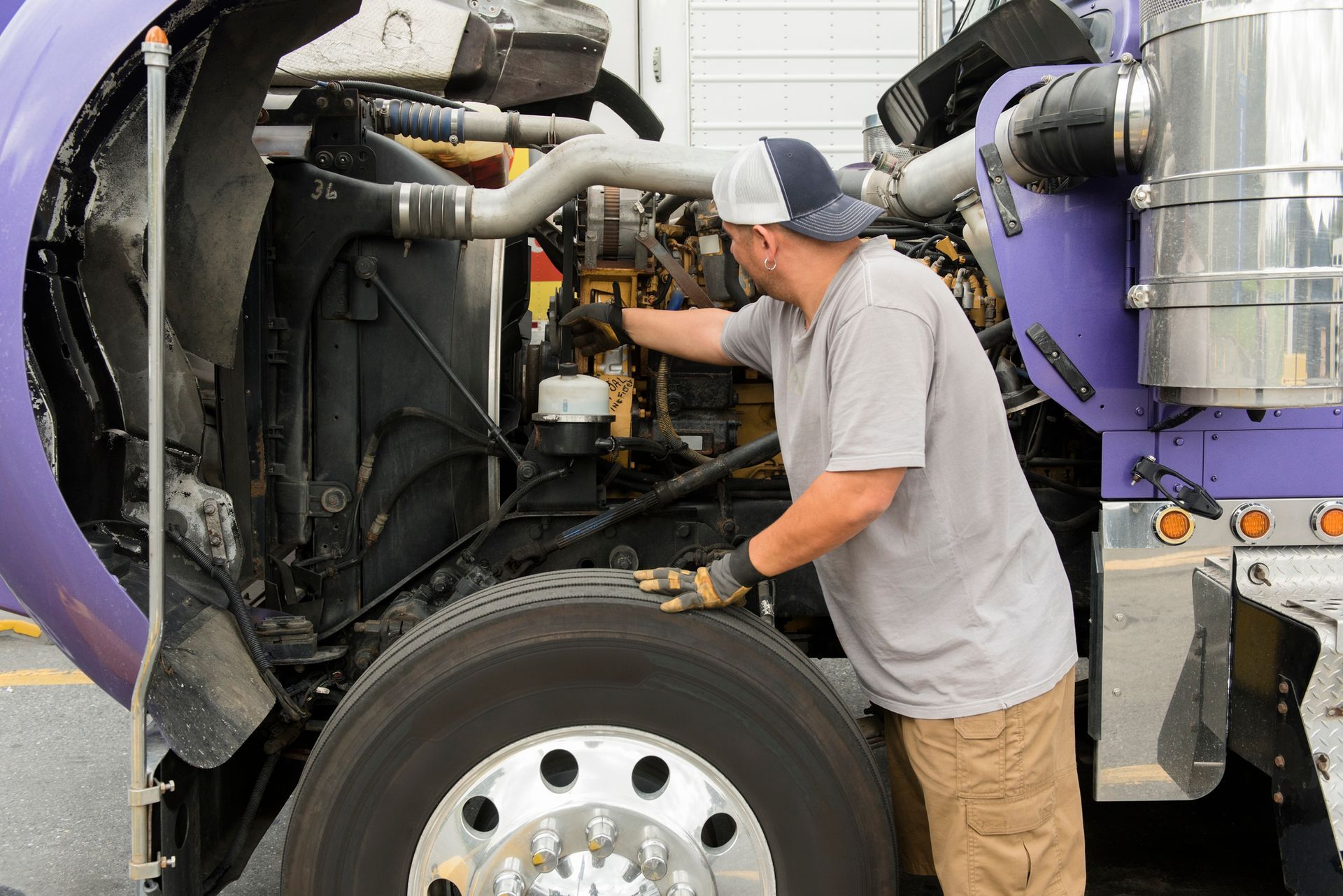 Mechanic works on a truck engine, wearing a hat and gloves, outdoors.