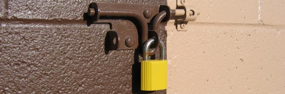A yellow padlock is locked through a brown metal sliding bolt attached to a textured door next to a light wall.