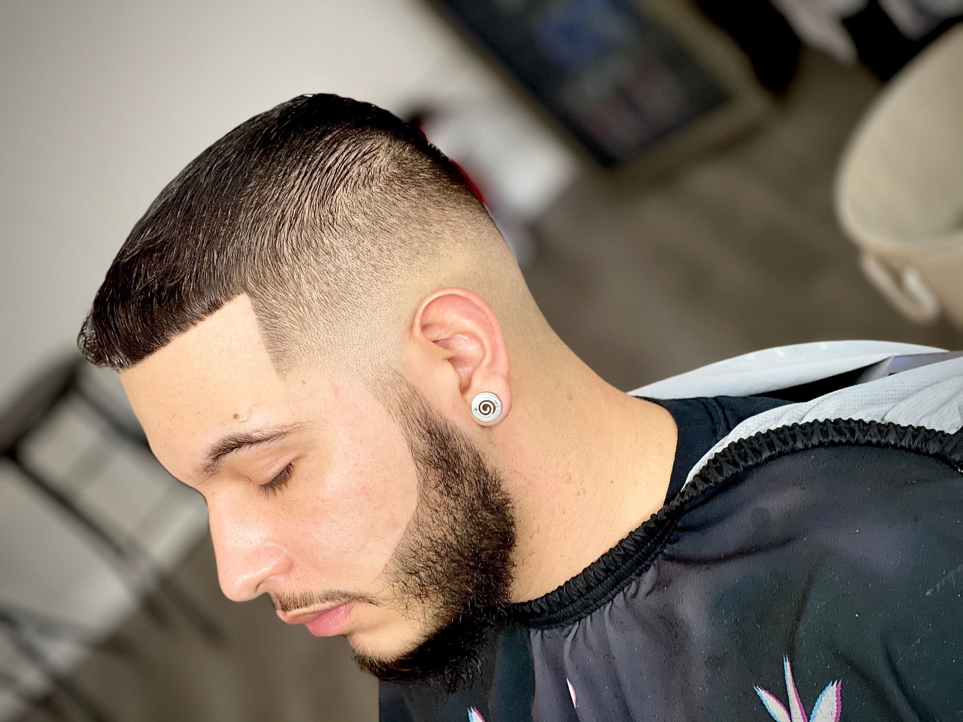 Man with a faded haircut and beard, wearing a hooded sweatshirt, eyes closed, in a barber setting.