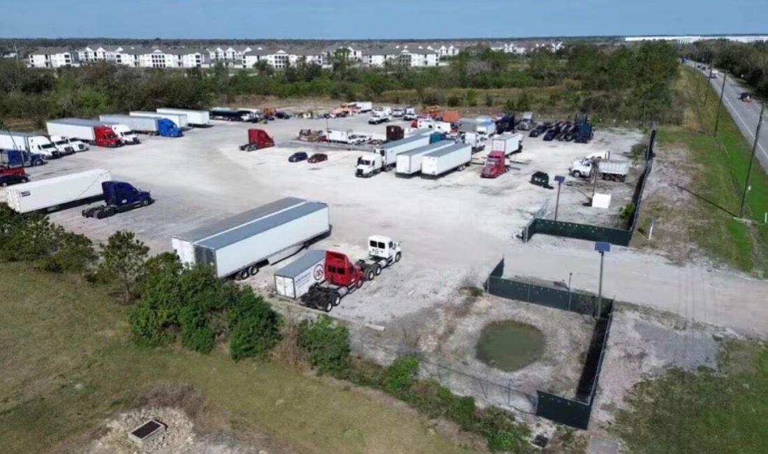 Aerial view of a truck stop with semi-trucks, trailers, and a small pond in a fenced area.