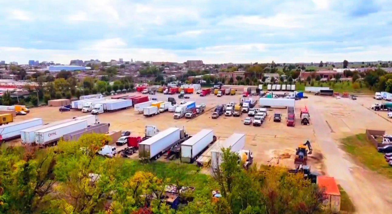 Trucks parked in a large lot, likely a trucking depot, under a cloudy sky.