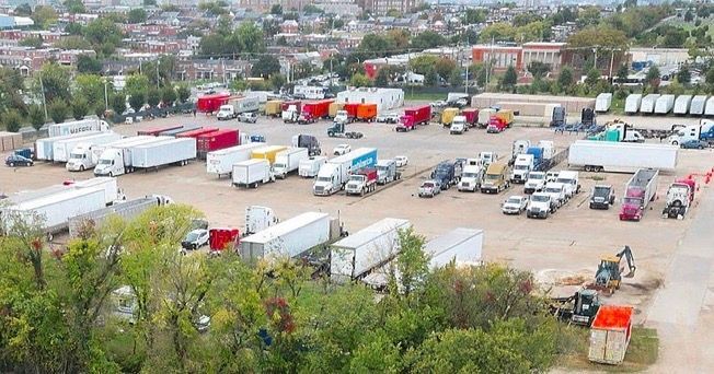 Trucks and trailers parked in an outdoor lot, near a town with buildings and trees.