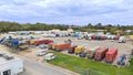 Truck yard with numerous semi-trucks, trailers, and shipping containers under cloudy sky.