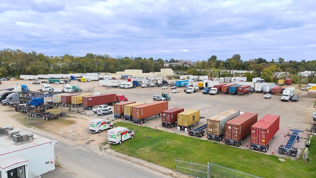 Truck yard with numerous semi-trucks, trailers, and shipping containers under cloudy sky.