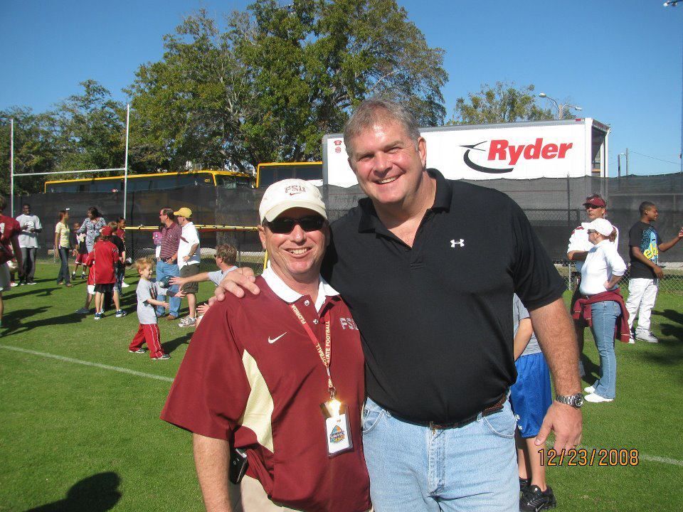 Macs arm around a tall man wearing a black shirt, posing outdoors on a green field. A Ryder truck is in the background.