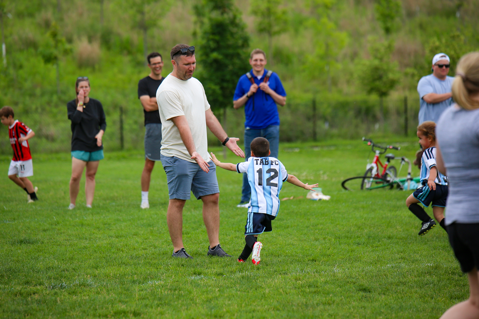 Soccer game on a grassy field, with a child in a striped jersey running toward an adult holding a ball.