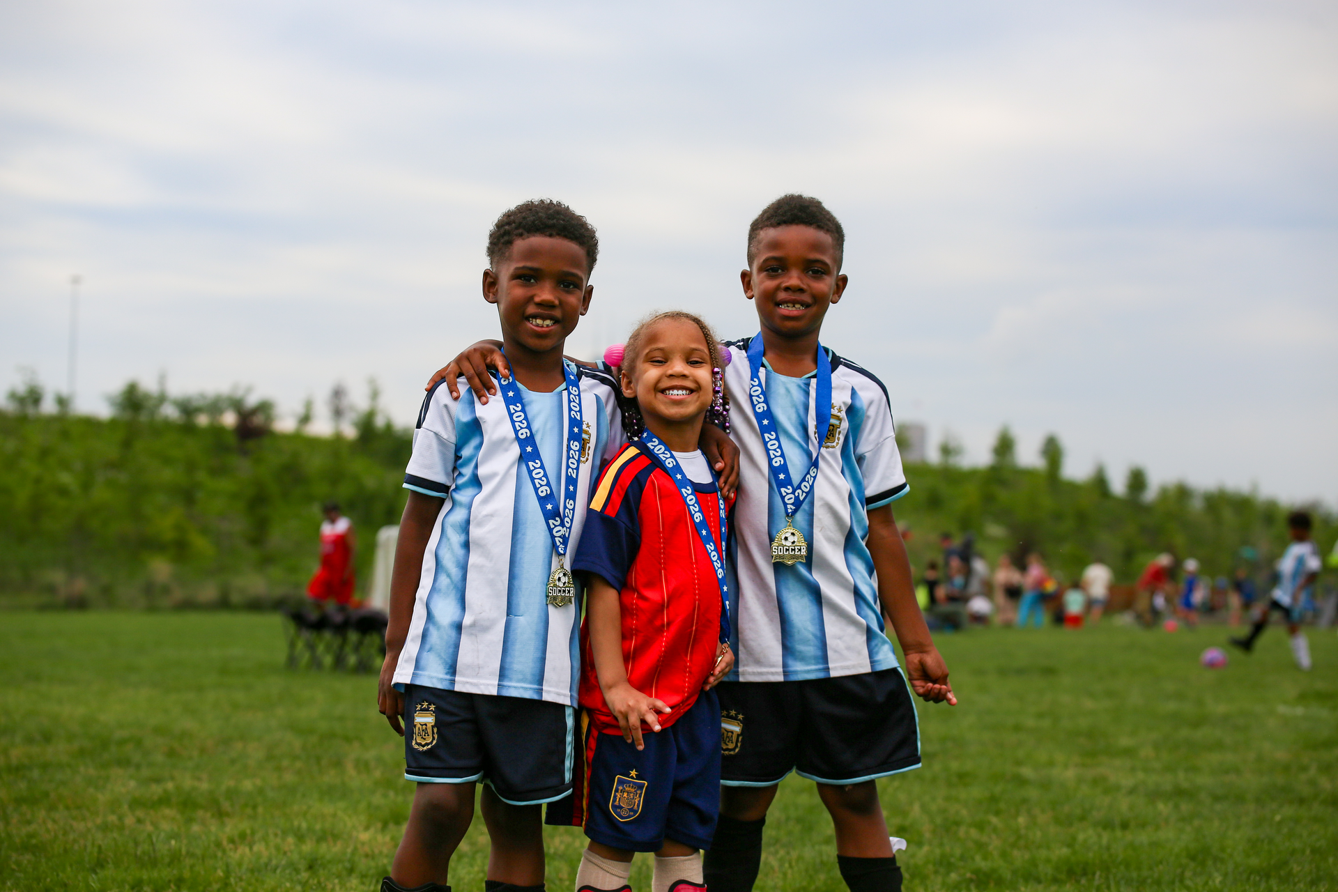 Three smiling children standing together on a grassy field, wearing blue jerseys and medals.