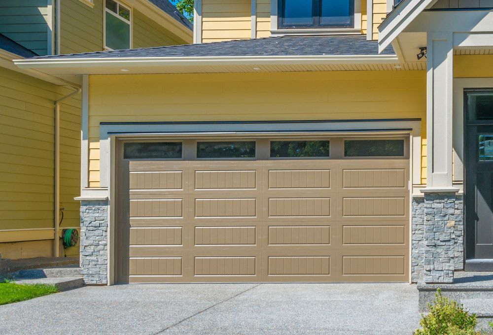 Tan garage door with windows above, on a yellow house. Concrete driveway.
