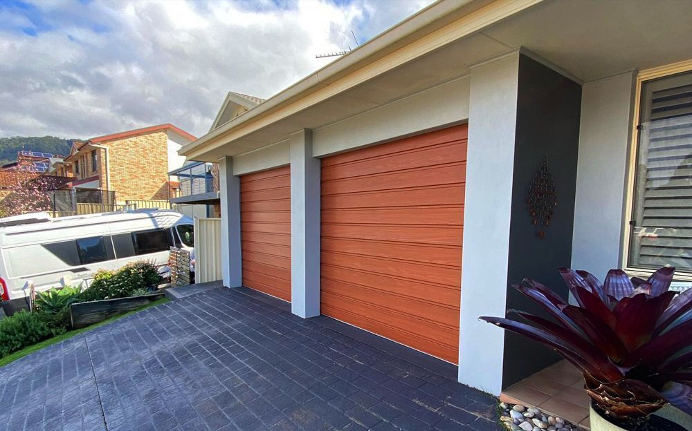 Two orange garage doors on a light grey building, dark grey driveway, with a red plant in the foreground.